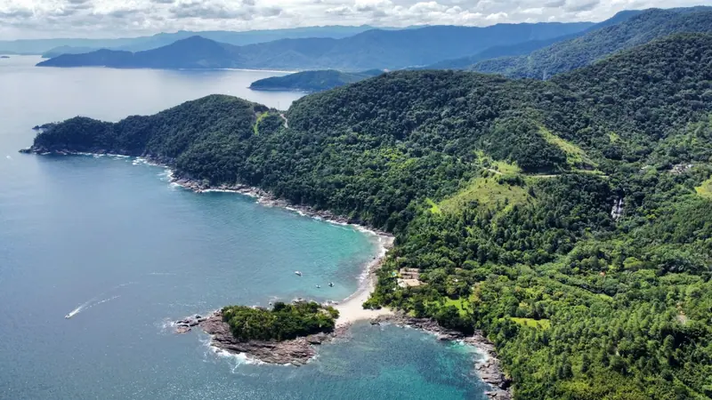 Vista aérea de Calhetas com a península, o costão de mata e a Cachoeira de Calhetas descendo o morro à direita