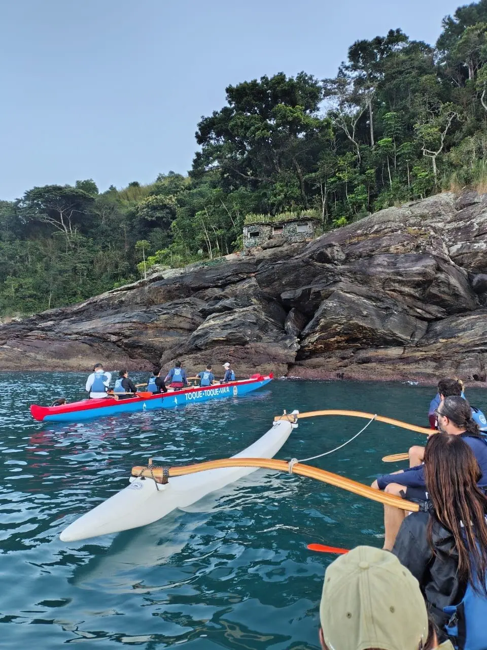 Canoa havaiana passando ao lado de um costão de pedra coberto de mata