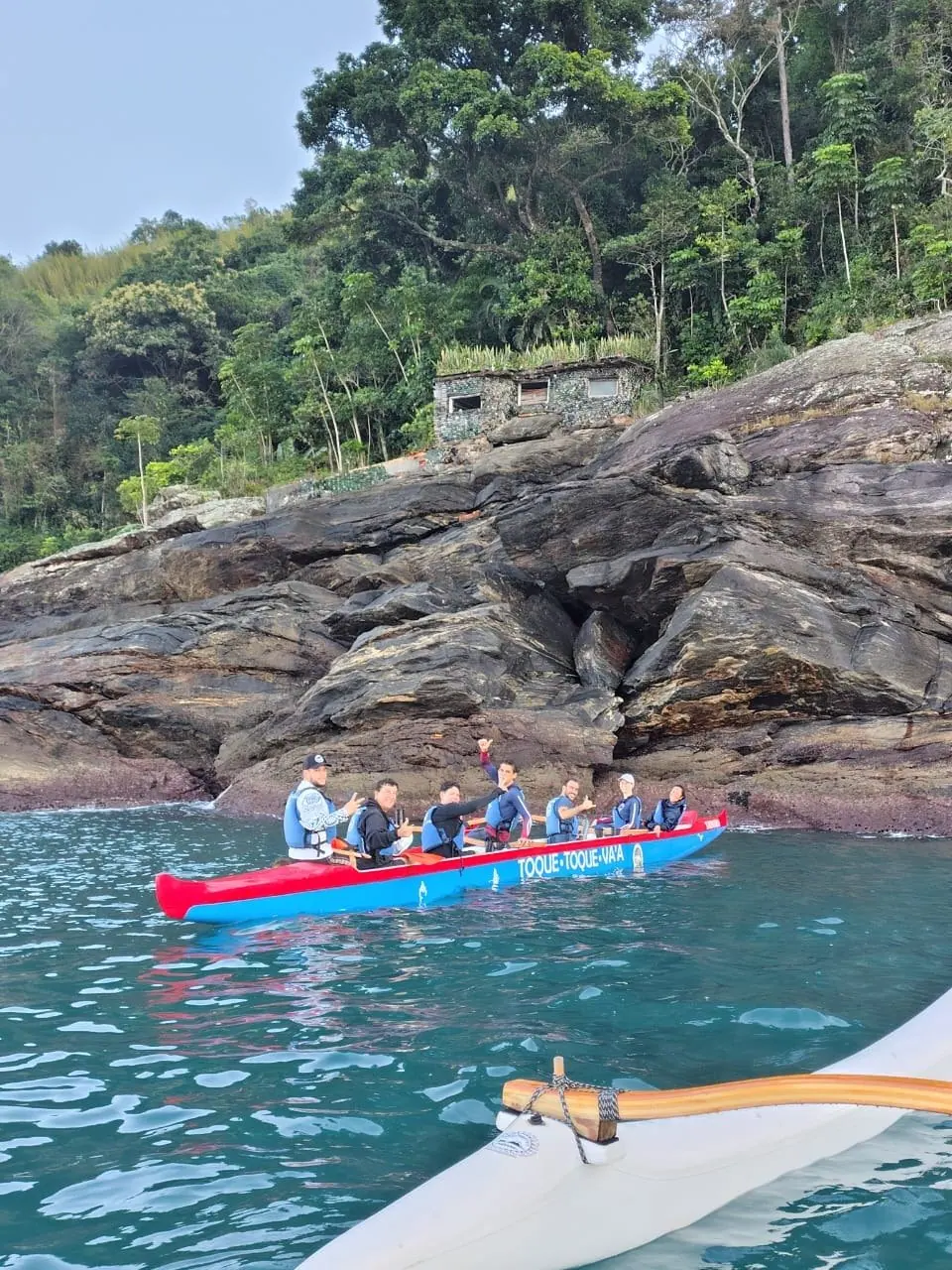 Canoa havaiana vermelha junto a um costão de pedra e mata atlântica