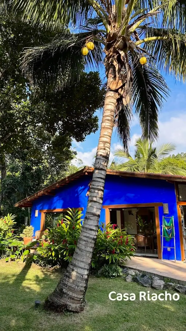Casa Riacho, de fachada azul, entre coqueiros e o jardim tropical da Vila